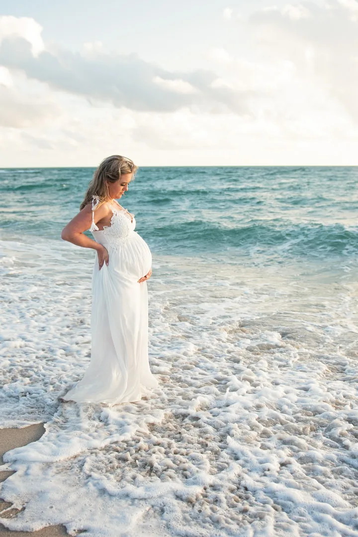 Meli B. Photography, a Miami-based natural light maternity photographer, captures the serene beauty of a pregnant woman in a white dress standing in the ocean.